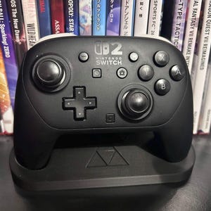 Puede incluir: Un mando Nintendo Switch Pro negro sobre una base de carga negra. El mando presenta un logotipo central de Nintendo Switch, dos joysticks, una cruceta y botones de acción estándar etiquetados como A, B, X e Y. La base del soporte tiene un diseño triangular en relieve.