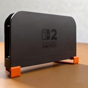 Pode incluir: Uma base de carregamento preta para consola Nintendo Switch 2 com o logótipo da Nintendo Switch e o número 2 impressos na frente. O dispositivo retangular é suportado por dois pés de plástico laranja brilhante nos cantos inferiores.