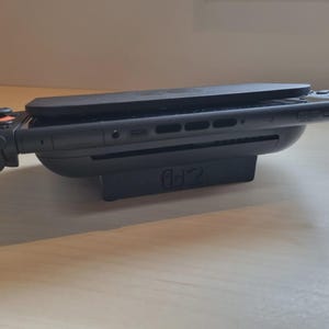 Peut inclure: Une console de jeu noire avec des manettes attachées, posée sur une station de charge noire. La console a un design élégant et moderne avec des boutons et des ports visibles. Les manettes ont des accents orange et noir.