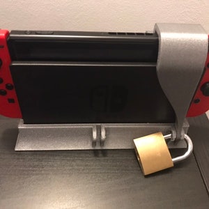 Puede incluir: Una consola Nintendo Switch asegurada en un soporte gris impreso en 3D con un candado de latón. La Switch tiene mandos rojos y está colocada sobre una superficie oscura.