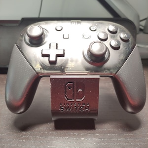 Peut inclure: Une manette noire Nintendo Switch est affichée sur un support noir avec les mots "Nintendo Switch" imprimés sur le devant. La manette est translucide et a une surface texturée.