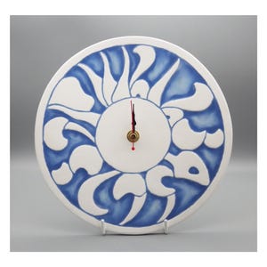 May include: A round, white ceramic clock with a blue floral pattern. The clock face has a central circle with a brass-coloured centre and red hands. The clock is displayed on a white stand.