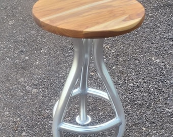 May include: A wooden stool with a round, oval-shaped seat and a silver metal base. The base has three legs that curve outward and then inward, creating a unique design.