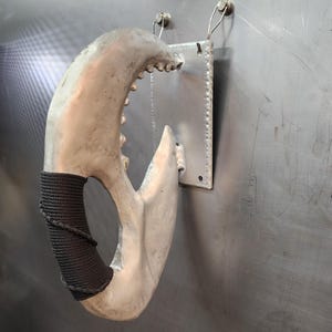 May include: A silver-colored metal claw with a black rope handle. The claw is mounted on a wall with two metal hooks.