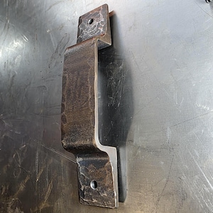 May include: A rustic, hand-forged metal door pull with a dark, textured finish. The handle features a rectangular grip and mounting plates with pre-drilled holes for easy installation. The metal has a hammered appearance, adding to its handcrafted aesthetic.