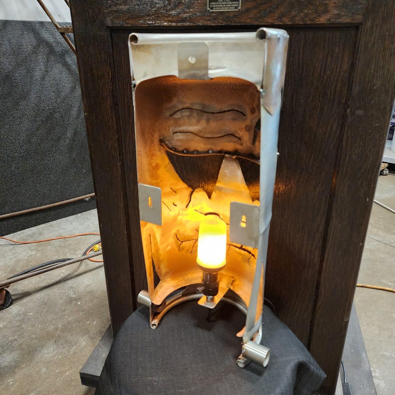 May include: A unique, handcrafted lamp with a metal frame and a warm, yellow light. The lamp's interior has a textured, rusted appearance, and it is mounted on a dark wooden structure. The design is industrial and artistic.