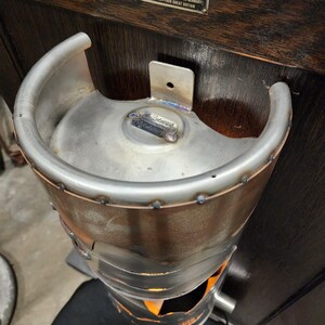 May include: A handcrafted metal fire pit made from a repurposed keg. The fire pit has a silver finish with welded seams and a curved handle. The bottom section has an opening for airflow, and the interior glows with the light of a fire.