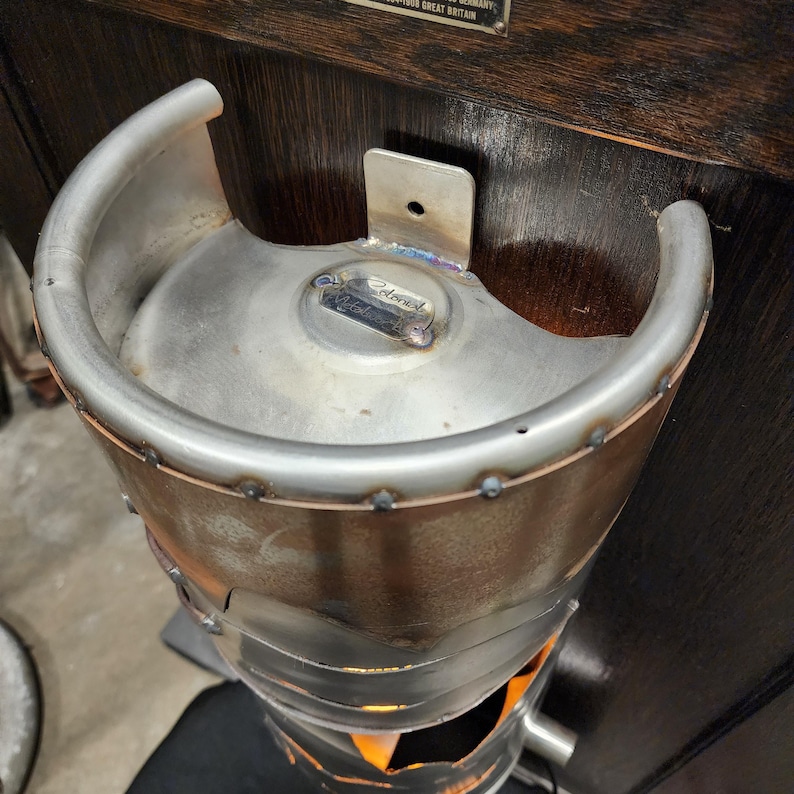 May include: A handcrafted metal fire pit made from a repurposed keg. The fire pit has a silver finish with welded seams and a curved handle. The bottom section has an opening for airflow, and the interior glows with the light of a fire.