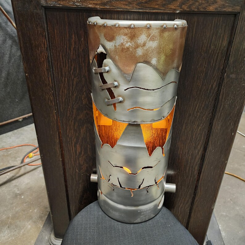 May include: A cylindrical metal lamp with a Frankenstein monster design. The lamp has a riveted top and a distressed, rusty finish. The face is cut out with stitched details and a glowing orange light inside. The lamp is approximately 12 inches tall.