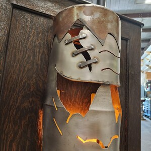 May include: A cylindrical metal lamp with a distressed, industrial aesthetic. The lamp features a riveted design with cut-out shapes, revealing a warm, orange glow from within. The top section has a jagged edge, adding to its unique character.