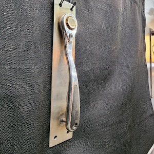 May include: A silver ratchet wrench attached to a rectangular metal plate. The wrench has a curved handle and a hexagonal head. The plate is mounted on a dark gray textured surface.