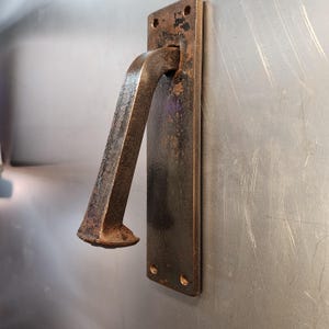 May include: A rustic, bronze-colored door handle with a rectangular backplate and a curved, angled handle. The handle is mounted on a metallic surface, showcasing a vintage aesthetic. The handle has a distressed finish.