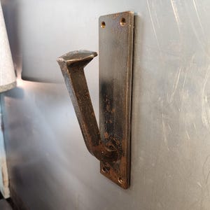 May include: A wall-mounted hook featuring a dark, weathered metal railroad spike attached to a rectangular metal backplate. The backplate has two circular screw holes at the top and bottom, and the entire piece displays a textured, aged finish.