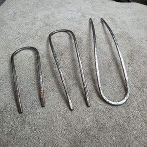 May include: Three handcrafted metal hair pins in varying sizes. The pins have a hammered texture and a silver finish. The U-shaped pins are designed for securing hair in place. The pins are displayed on a neutral-colored surface.