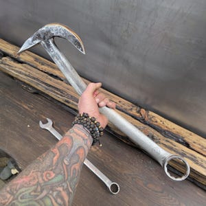 May include: A large, custom-made metal tool with a curved, pointed end and a wrench-like base. The tool is held in a hand, with a smaller wrench visible below. The metal has a distressed, industrial look.