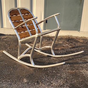 May include: A modern rocking chair with a polished metal frame and a backrest made of wooden slats. The chair has curved rockers and armrests, offering a sleek and comfortable design. The chair is designed for outdoor or indoor use.