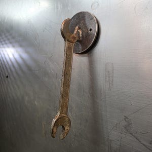 May include: A rusty, vintage wrench attached to a circular metal plate. The wrench is a light brown color and shows signs of wear. The plate is dark brown with a single hole. The items are mounted on a gray metal surface.