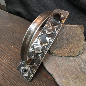 May include: A rustic, metal door pull with a textured, square pattern on the base plate and a curved handle. The metal has a dark, aged finish, suggesting a handcrafted or vintage aesthetic. The door pull is designed for functionality and adds a touch of industrial charm.