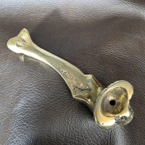 May include: A vintage brass drawer pull with a unique, ornate design. The pull features a long, curved handle with a decorative end and a circular base with a central hole. The brass has a slightly aged appearance.