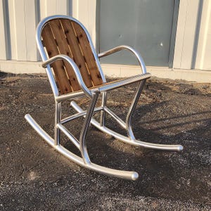 May include: A modern rocking chair with a polished metal frame and wooden seat and back. The chair features curved armrests and rockers, offering a comfortable and stylish seating option. The wooden back is made of vertical wooden planks.