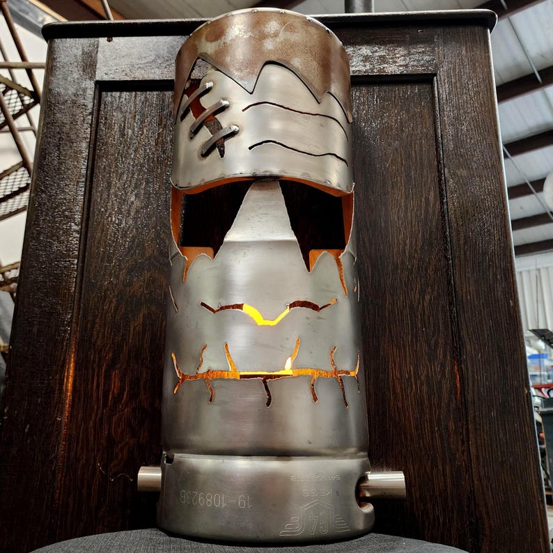 May include: A handcrafted metal lamp with a Frankenstein-like design. The lamp features cut-out shapes for eyes and mouth, with stitched details. The top has a rusted finish, and the interior glows with warm light, creating a unique decorative piece.