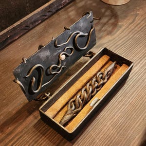 May include: A black metal box with a hinged lid decorated with a swirling design. The lid is open, revealing a brown leather-lined interior with a long, twisted metal object inside.
