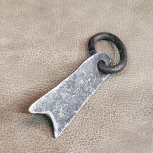 May include: A rustic, hand-forged metal key chain with a simple, flat design. The key chain has a loop at the top and a pointed end. The metal has a dark, aged finish.
