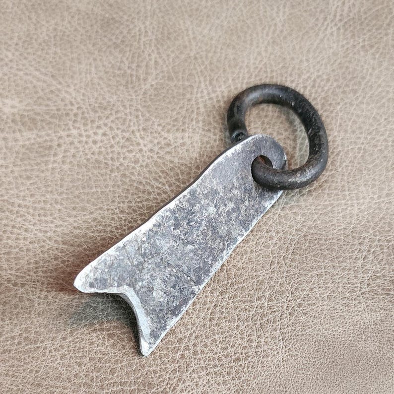 May include: A rustic, hand-forged metal key chain with a simple, flat design. The key chain has a loop at the top and a pointed end. The metal has a dark, aged finish.