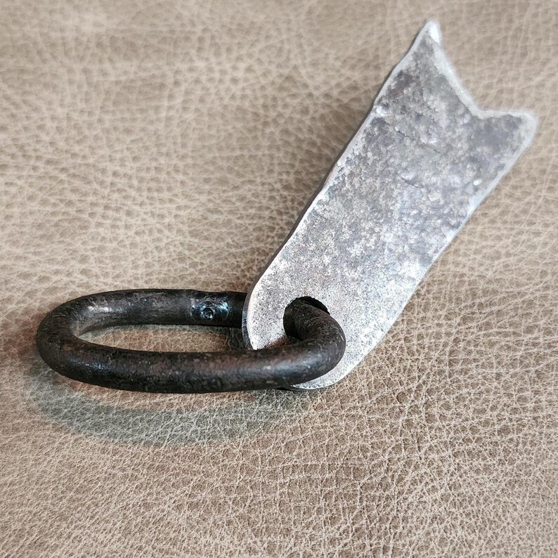 May include: A rustic, hand-forged metal door handle with a simple, oval ring and a flat, rectangular plate. The handle is made of dark metal with a rough, hammered finish.
