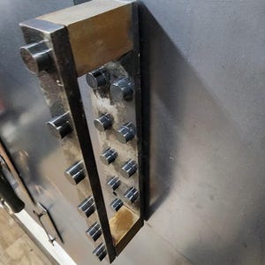 May include: A metal door handle with a vertical row of round black knobs. The handle is attached to a wall with a metal plate.