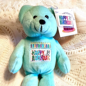 May include: A teal plush teddy bear with a black nose and embroidered "Happy 37th Birthday" text. The bear has a tag that reads "Happy Birthday" with a cake graphic. A cute and cuddly birthday gift.