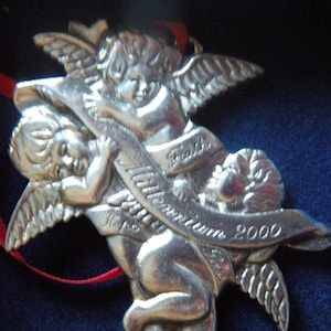 May include: Silver Christmas ornament featuring three angels with wings. The angels are holding a banner that reads "Faith, Hope, Love 2000".
