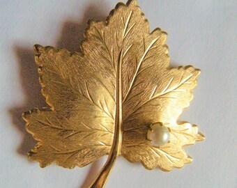 Op de afbeelding: Goudkleurige broche in de vorm van een blad met gedetailleerde nerven en een getextureerd oppervlak. Een kleine, ronde, crèmekleurige parel is op het blad geplaatst. De broche is waarschijnlijk van metaal en is een decoratief accessoire.