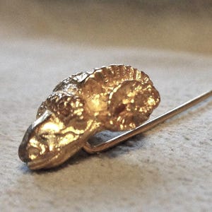 May include: A gold-colored stick pin featuring a ram's head design. The ram's head is detailed with textured fur and horns. The pin has a long, slender shaft with a small, rounded end. This jewelry item is likely a vintage or antique accessory.