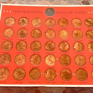 Può includere: Una carta rossa e bianca con il titolo "Presidential Hall of Fame" e 39 gettoni rotondi color rame con i volti dei presidenti degli Stati Uniti.