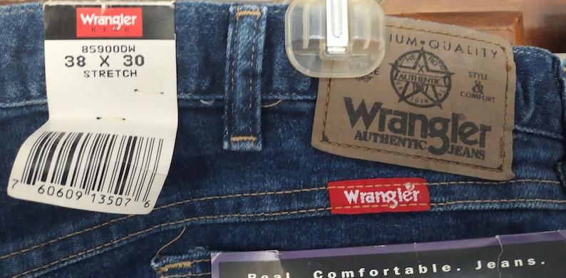 Puede incluir: Un par de pantalones vaqueros azules con una etiqueta blanca que dice "Wrangler" y "85900DW 38 X 30 STRETCH" y una etiqueta marr&oacute;n que dice "Wrangler Authentic Jeans".