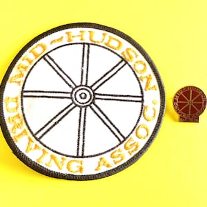 May include: A white and black embroidered patch and a pin with the text "MID-HUDSON DRIVING ASSOC." and a wheel design. The patch has a black border and yellow text. The pin is red and gold with the text "EST. 1974".