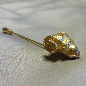 May include: A gold-toned tie pin featuring a ram's head design. The ram's head has detailed horns and a textured surface, with blue accents. The pin is set against a neutral background.