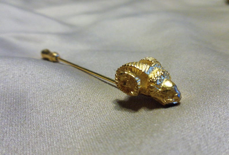 May include: A gold-toned tie pin featuring a ram's head design. The ram's head has detailed horns and a textured surface, with blue accents. The pin is set against a neutral background.