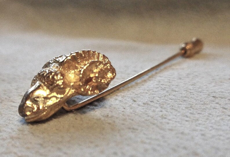 May include: A gold-colored stick pin featuring a ram's head design. The ram's head is detailed with textured fur and horns. The pin has a long, slender shaft with a small, rounded end. This jewelry item is likely a vintage or antique accessory.