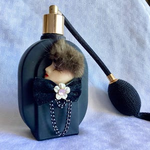 Puede incluir: Botella de perfume de vidrio negro con un retrato en blanco y negro de una mujer con un sombrero de piel y un lazo negro. La botella tiene un atomizador dorado y una borla negra.
