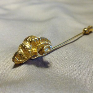 May include: A gold-colored stick pin featuring a ram's head design. The ram's head is detailed with textured fur and horns. The pin is made of a gold-toned metal and is set against a neutral background.