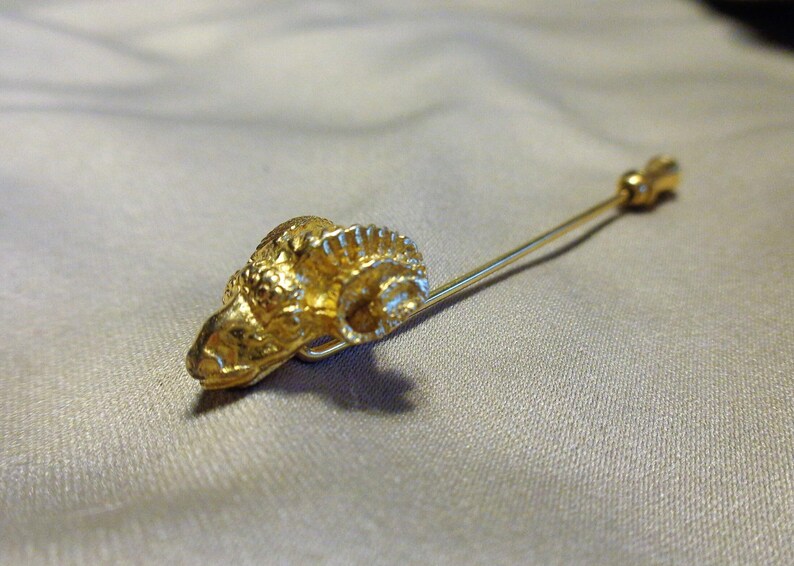 May include: A gold-colored stick pin featuring a ram's head design. The ram's head is detailed with textured fur and horns. The pin is made of a gold-toned metal and is set against a neutral background.