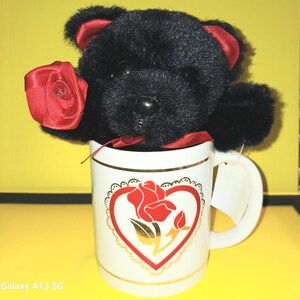Könnte beinhalten: Ein schwarzer Teddybär, der eine rote Rose hält, sitzt in einer weißen Keramiktasse mit goldener Verzierung und einem roten Rosenmuster.