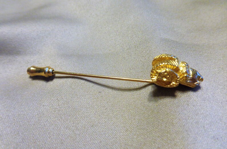 May include: A gold-colored stick pin featuring a detailed ram's head design. The pin has a long, slender shaft and a decorative, rounded end. The ram's head is intricately sculpted, showcasing the horns and facial features. The pin is set against a neutral background.