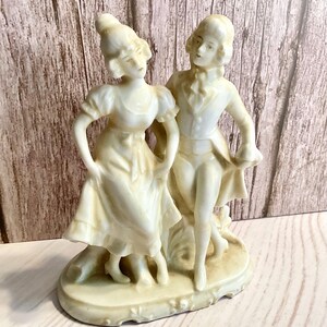 May include: A porcelain figurine of a couple dancing. The woman is wearing a long dress and the man is wearing a suit. The figurine is painted in a cream color.