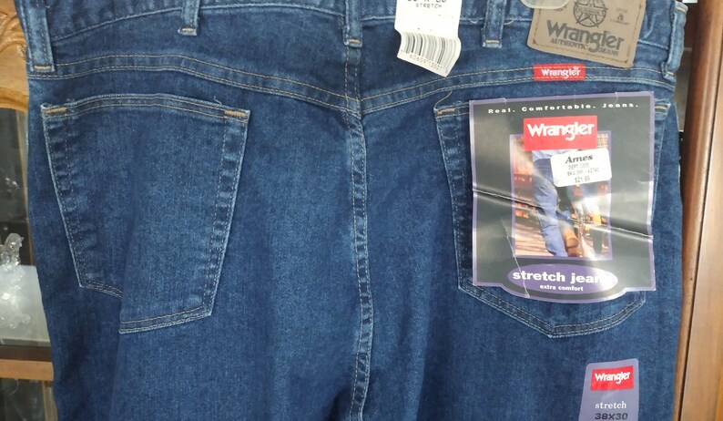 Puede incluir: Un par de pantalones vaqueros azules con un bolsillo trasero y una etiqueta de Wrangler adjunta. La etiqueta dice "Real. Comfortable. Jeans." y "Stretch Jeans Extra Comfort". Los vaqueros son talla 38x30.