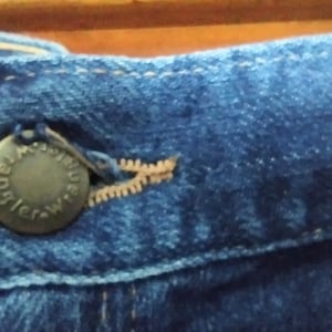 Puede incluir: Primer plano de una cintura de mezclilla azul con un bot&oacute;n de metal que dice "Wrangler" en un dise&ntilde;o circular. El bot&oacute;n est&aacute; unido a una cremallera.