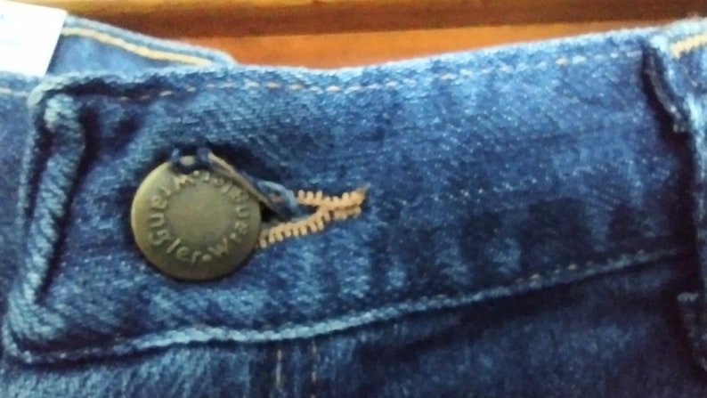 Puede incluir: Primer plano de una cintura de mezclilla azul con un bot&oacute;n de metal que dice "Wrangler" en un dise&ntilde;o circular. El bot&oacute;n est&aacute; unido a una cremallera.