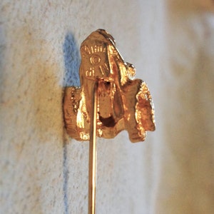 May include: A gold-colored lapel pin with a detailed, textured design. The pin features a stylized shape with visible text. The pin has a long, thin, gold-colored pin backing.